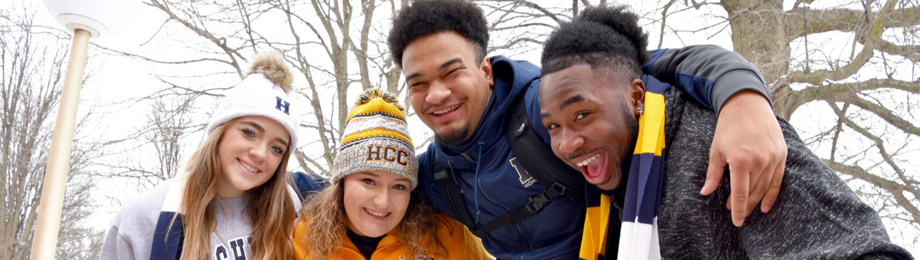Four students standing in the snow, huddle in a group together. They are wearing HCC hats and gloves.