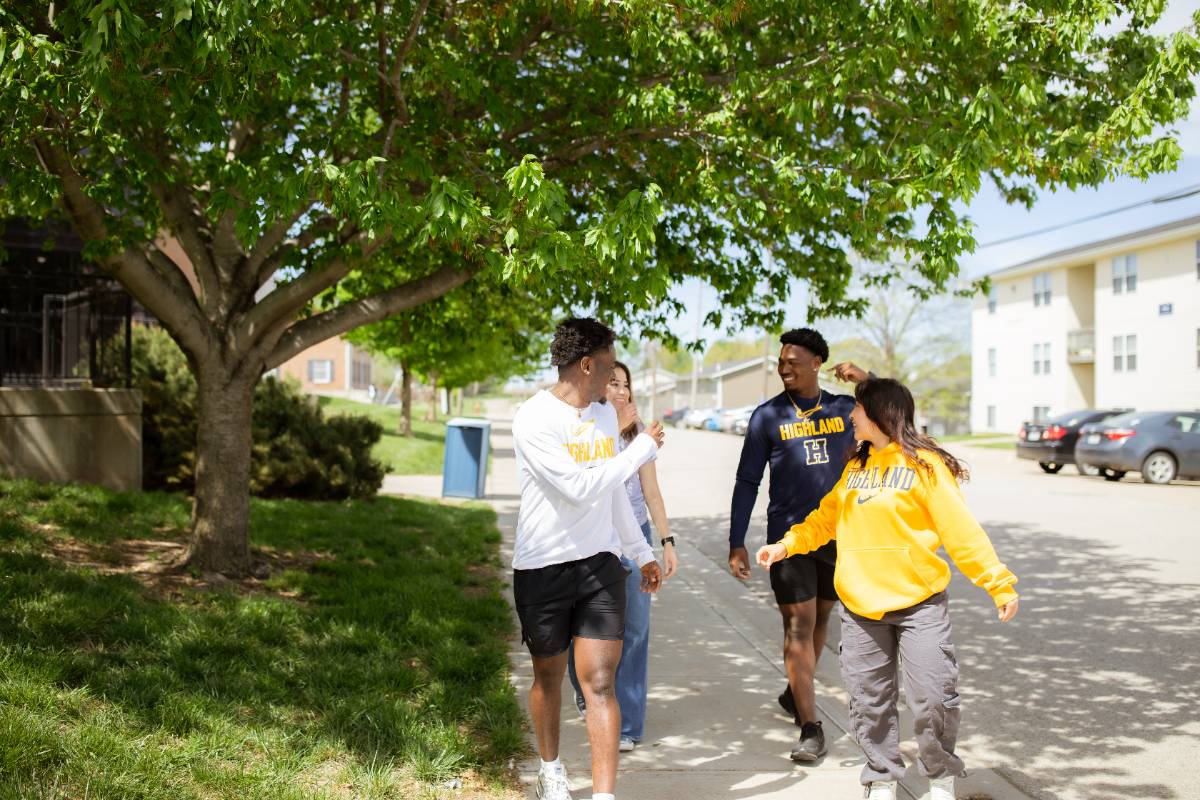 Group of four students walking on the sidewalk together, turning to look toward each other as they walk. 