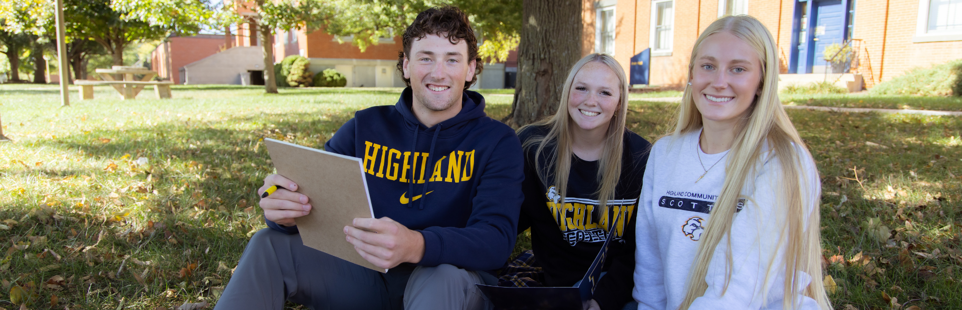 Three students on the lawn, smiling at the camera. They are wearing blue and white sweatshirts with "Highland" on them.