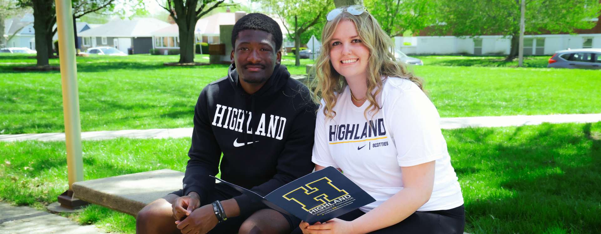 Two students sitting on a bench outside, smiling toward the camera while waring Highland apparel.