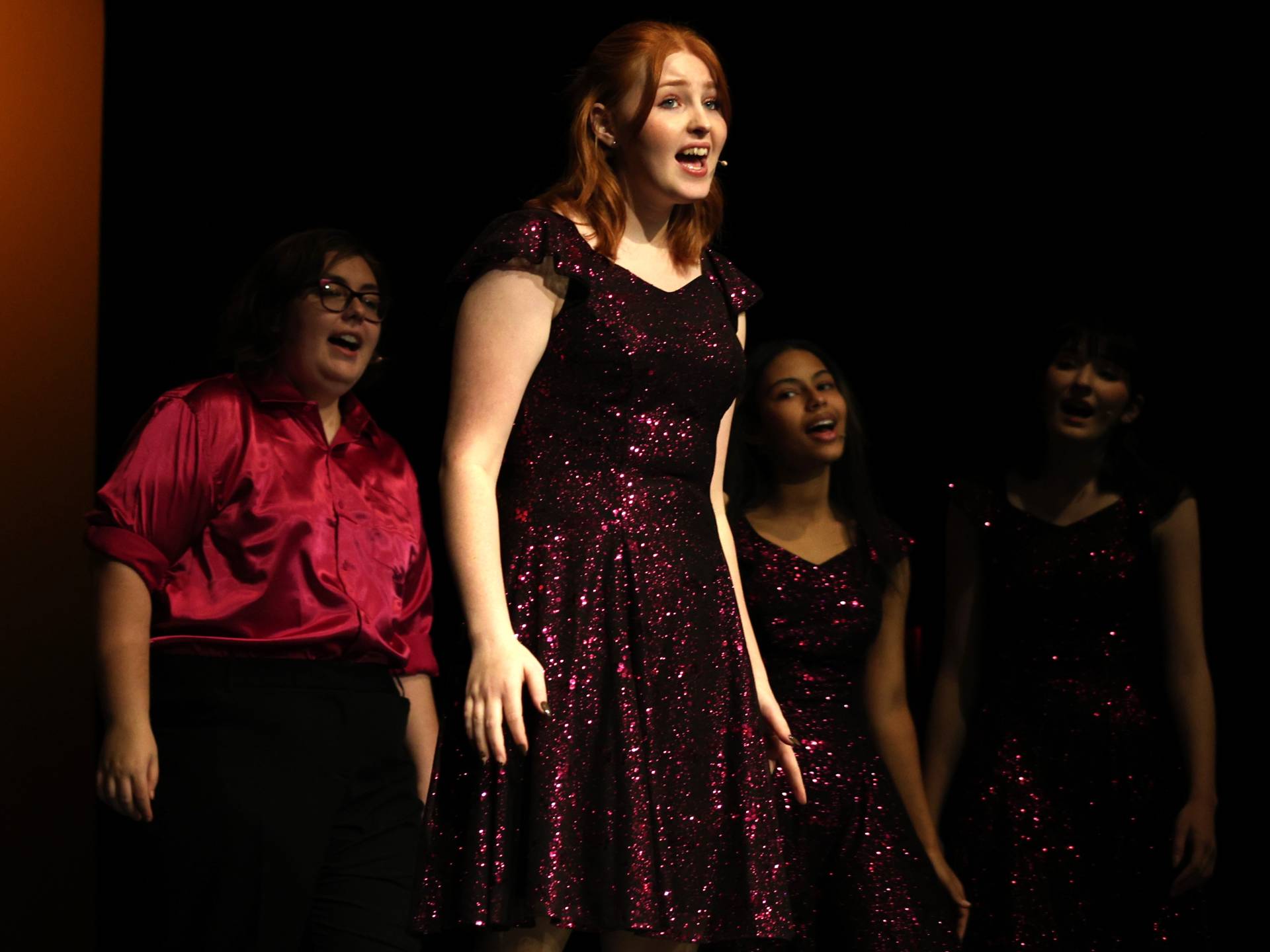 A student singing in the spotlight with three choir members singing behind her