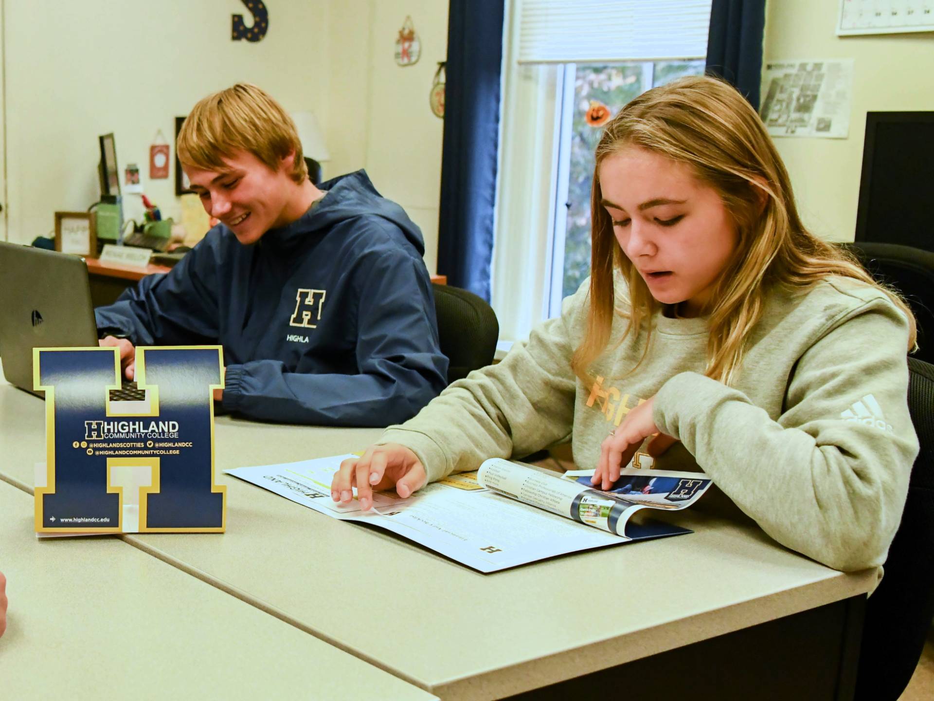 Two students at a table together, one is reading and the other is on a laptop.