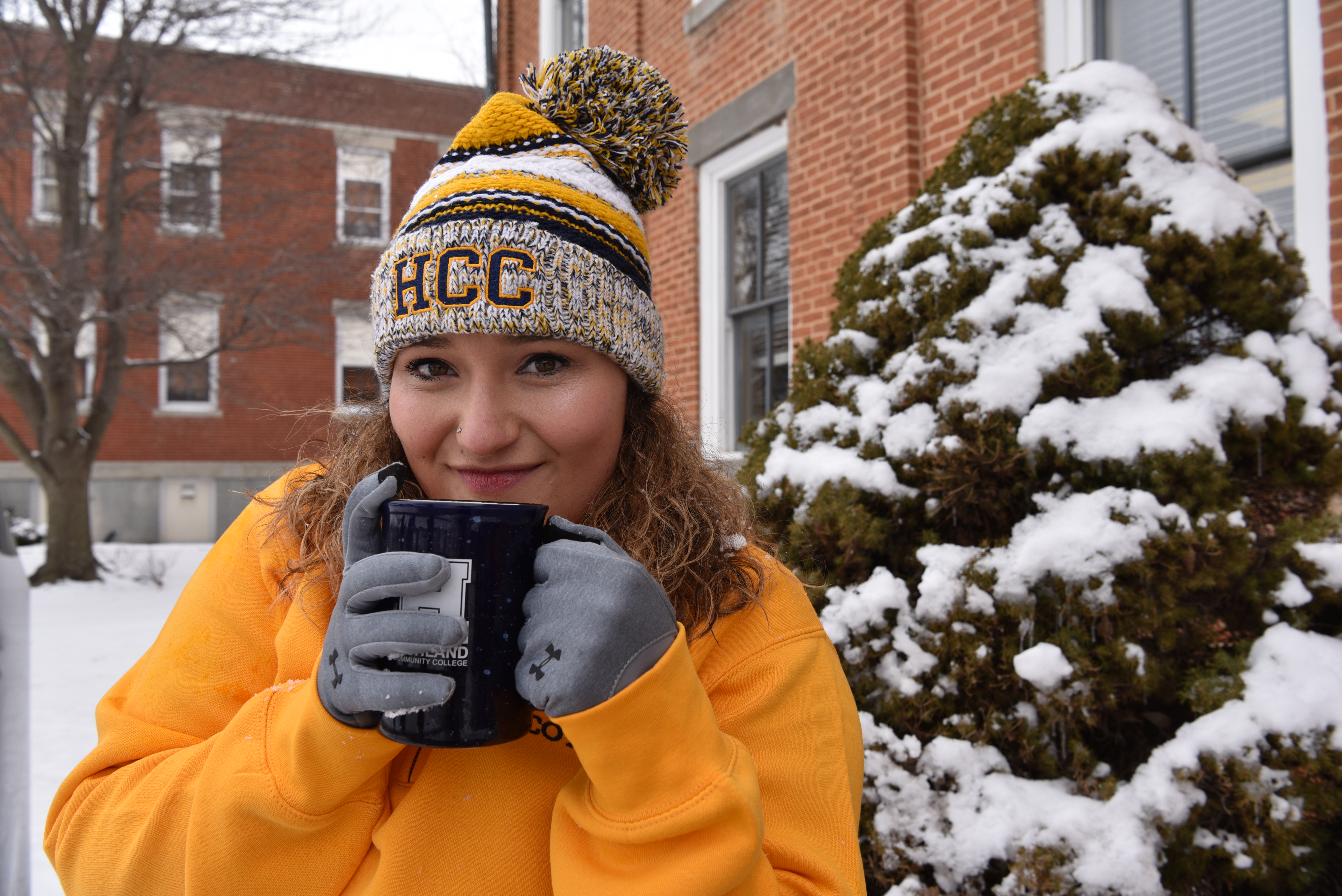 A smiling person in a yellow sweatshirt and winter hat holds a mug with both hands outside in the snow, standing near a snow-covered bush and brick buildings.